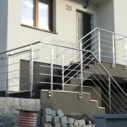 Zewnętrzne schody z nowoczesną balustradą nierdzewną, prowadzące do wejścia domu z numerem 20, widok z dołu na jasną elewację.