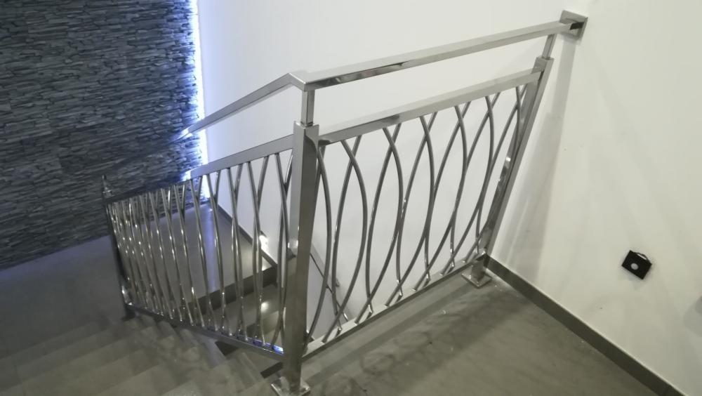 Nowoczesna balustrada ze stali nierdzewnej z dekoracyjnym wypełnieniem w kształcie krzyżujących się łuków, zamontowana przy schodach wewnątrz budynku z akcentem kamiennej ściany w tle.