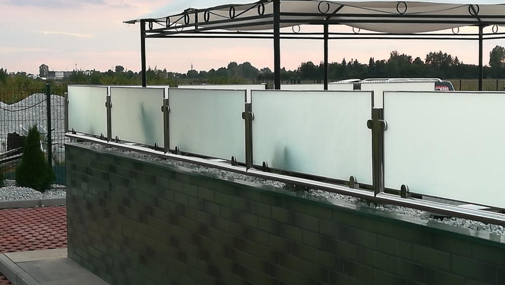 Szklana balustrada z nierdzewnymi słupkami na murku z cegły klinkierowej, widok na ogród i zadaszenie tarasu w tle.