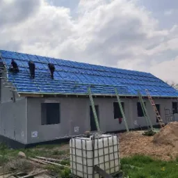 Budowa domu: trzy osoby pracują na dachu pokrytym niebieską folią, widoczne rusztowania i materiały budowlane na ziemi, ściany domu w surowym stanie z widocznymi oznaczeniami.