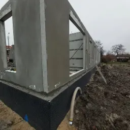 Surowy stan domu z prefabrykatów betonowych, widoczne fundamenty zabezpieczone czarną folią hydroizolacyjną, wystające rury, drabina oparta o ścianę, pochmurne niebo.