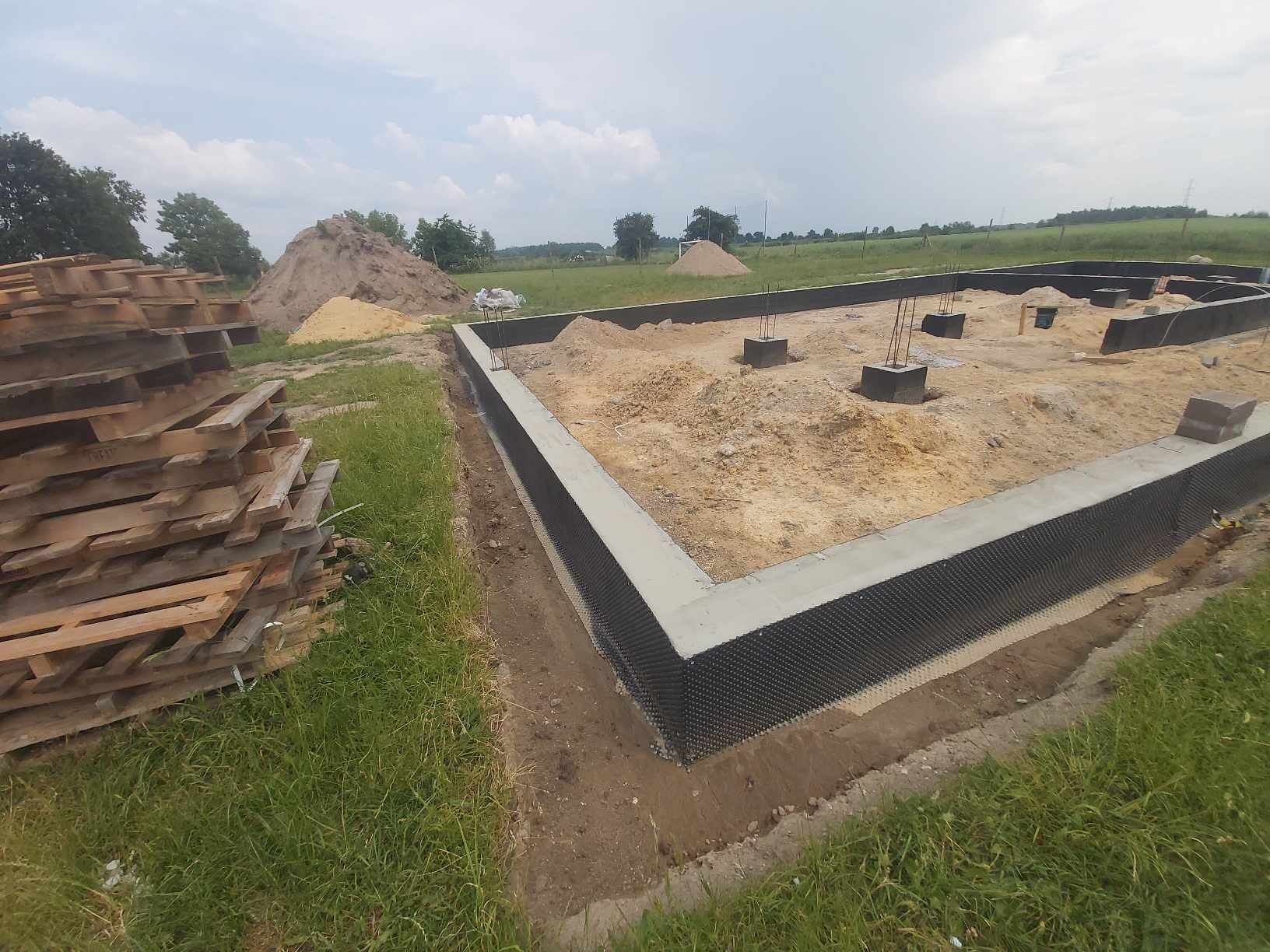Wylany fundament z hydroizolacją pionową w postaci folii kubełkowej, z wystającymi prętami zbrojeniowymi, widoczny stos palet w tle oraz usypana ziemia.