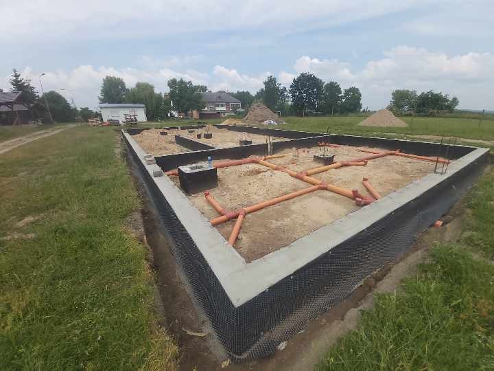 Wykop pod fundamenty z wykonaną izolacją pionową, z widocznymi rurami kanalizacyjnymi wewnątrz obrysu i wystającymi prętami zbrojeniowymi.