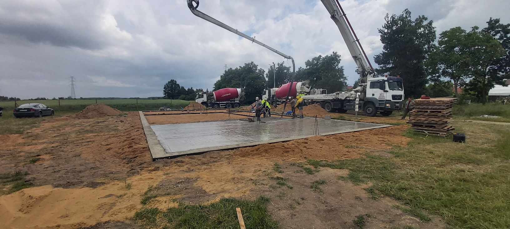 Wylewanie fundamentów pod budowę domu: ekipa budowlana wyrównuje świeży beton na płycie fundamentowej, widoczne dwa betonowozy z pompami, w tle słupy wysokiego napięcia.