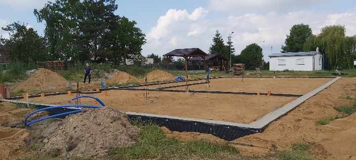 Wylane fundamenty pod budowę domu, widoczne zbrojenia i izolacja pionowa, na drugim planie sterta cegieł i pracownik budowlany.