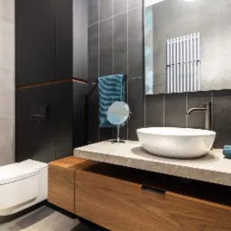 Nowoczesna łazienka z umywalką nablatową, drewnianą szafką pod umywalką, szarą glazurą ścienną i wiszącą toaletą.