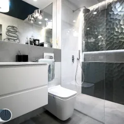 Nowoczesna łazienka z umywalką nablatową, podwieszaną toaletą i ścianą z dekoracyjnych, trójwymiarowych płytek w odcieniach szarości, oświetlonych listwą LED.