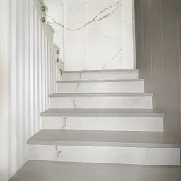 Nowoczesne schody z białego kamienia z szarymi żyłami, szklana balustrada, ściana w pionowe pasy i logo firmy w rogu.