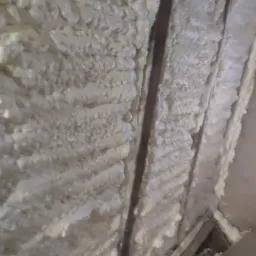 Poddasze wypełnione białą pianą izolacyjną między drewnianymi belkami, widoczne niedoskonałości w aplikacji i nierównomierna struktura piany.