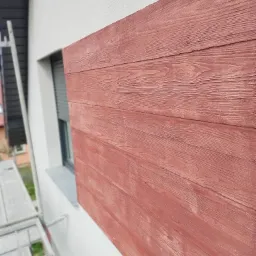 Fragment elewacji budynku z wykończeniem imitującym drewniane deski w odcieniach czerwieni, widoczne rusztowanie w tle, okno z opuszczonymi roletami.