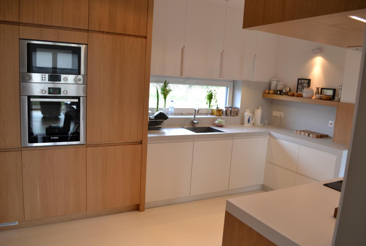 Nowoczesna kuchnia z drewnianymi frontami szafek, zabudowanym piekarnikiem i mikrofalówką, oraz białymi blatami i szafkami pod oknem z widokiem na zieleń.