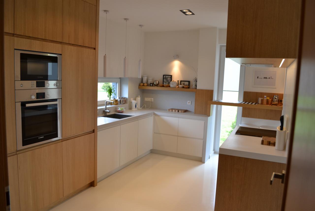 Jasna kuchnia z drewnianymi i białymi frontami, zabudowany piekarnik i mikrofalówka, szara ściana, drewniana półka z dekoracjami, okno z widokiem na zieleń.