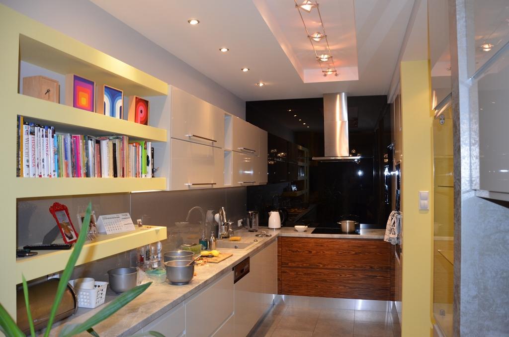 Nowoczesna kuchnia z jasnymi szafkami, drewnianym blatem, półkami z książkami i błyszczącą czarną ścianą, oświetlona punktowymi lampami sufitowymi.