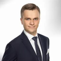 Portret mężczyzny w granatowym garniturze i krawacie w kropki, z białą poszetką w kieszeni, na jasnym tle. Uśmiech, kontakt wzrokowy.