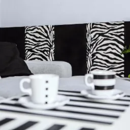 Aranżacja wnętrza z szarą sofą, poduszkami w kolorze białym i czarnym, panelem zagłówka z motywem zebry, dwoma filiżankami na stoliku w czarno-białe pasy oraz zieloną rośliną doniczkową.