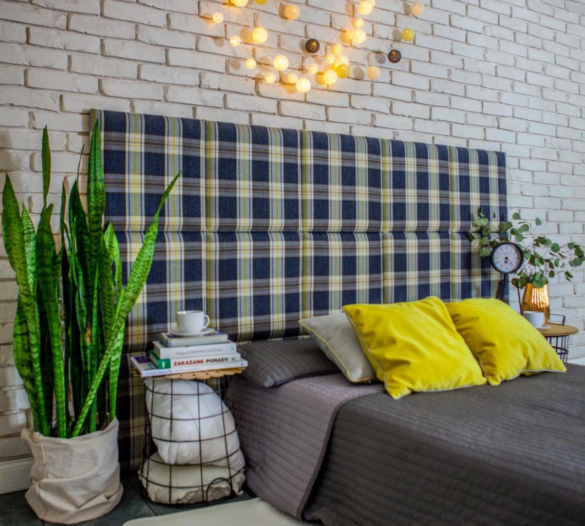 Tapicerowane wezgłowie łóżka w kratę, dopasowane kolorystycznie do zasłon, z dekoracjami: poduszkami, lampkami i roślinami, na tle ściany z białej cegły.