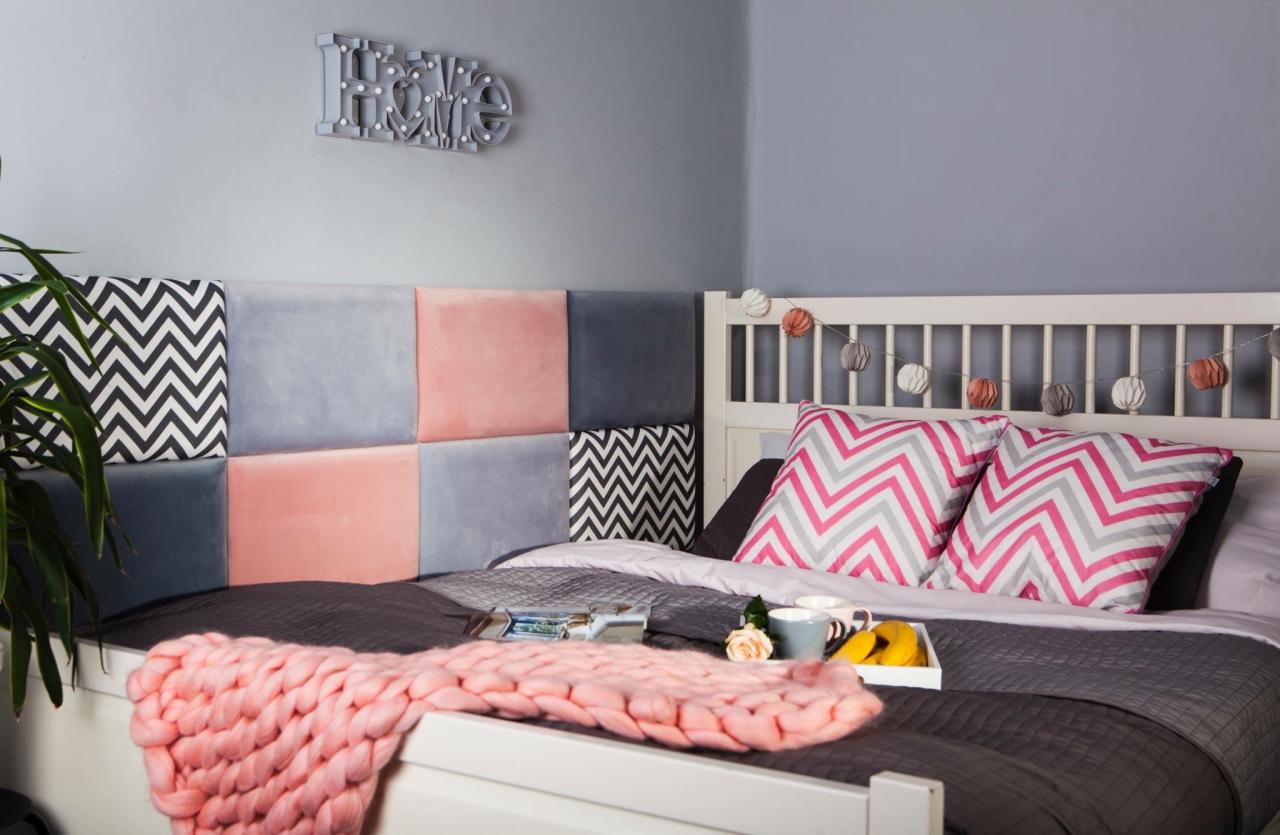 Tapicerowany panel zagłówka łóżka w odcieniach różu, szarości i czerni z geometrycznym wzorem, dekoracja ścienna HOME, śniadanie na tacy i pleciony koc na łóżku.