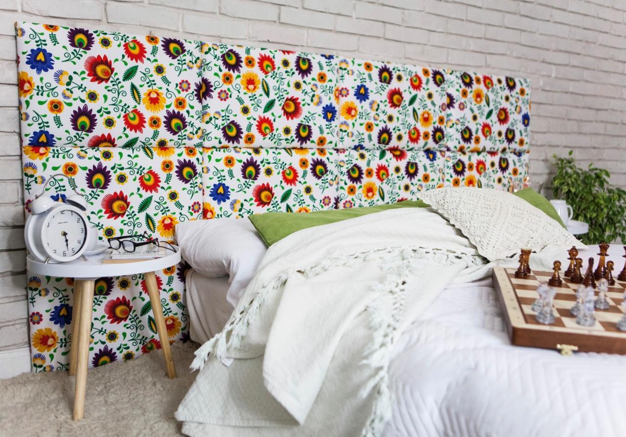 Tapicerowane wezgłowie łóżka w ludowe kwiaty, biała cegła w tle, stolik nocny z budzikiem i okularami, szachownica na pościeli.