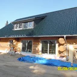 Dom w trakcie budowy z bali drewnianych, widoczne surowe drewno, okna bez wykończenia, niebieska plandeka na ziemi, dach pokryty ciemną dachówką.