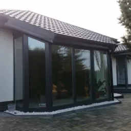 Nowoczesny dom z dużymi oknami PCV w czarnych ramach, widok z zewnątrz na kostkę brukową i ogród, z widoczną rośliną doniczkową wewnątrz.