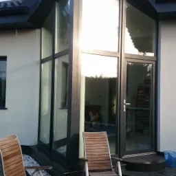 Nowoczesna bryła domu z dużym, przeszklonym narożnikiem i drzwiami wejściowymi, dwa drewniane krzesła na tarasie z kostki brukowej.