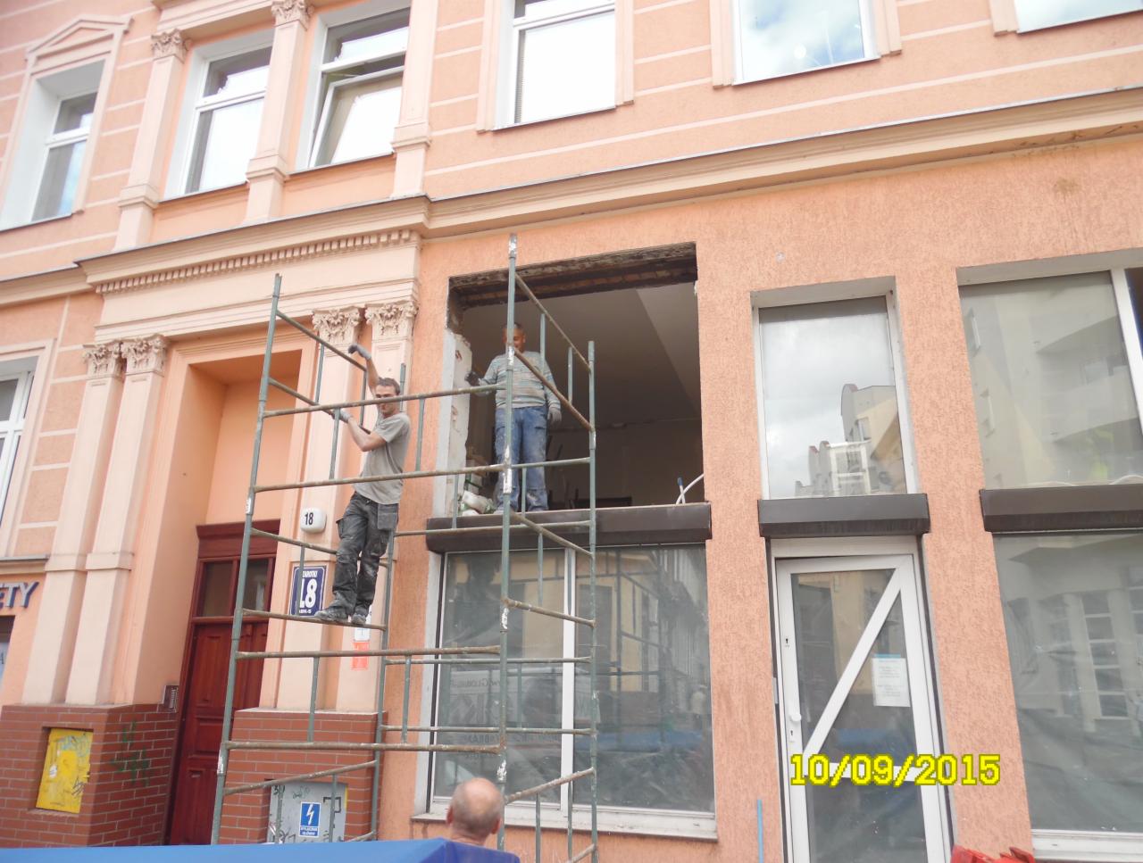 Dwóch monterów na rusztowaniu podczas wymiany okna w kamienicy, widoczne przygotowane otwory okienne i okno zabezpieczone folią.