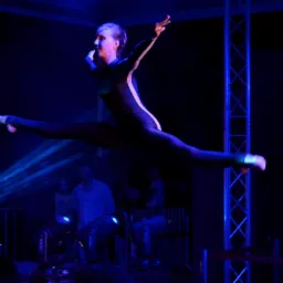 Kobieta w czarnym kostiumie z białym paskiem wykonuje szpagat w powietrzu na scenie oświetlonej niebieskim światłem, widoczny fragment konstrukcji scenicznej i reflektory