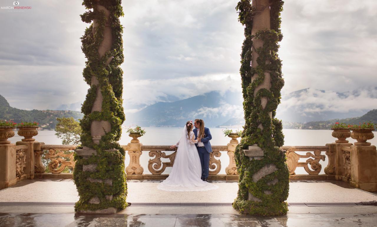 Para młoda całuje się na balkonie z widokiem na jezioro i góry, otoczona kolumnami porośniętymi bluszczem.