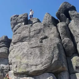 Dwóch mężczyzn w górach, jeden na szczycie formacji skalnej, drugi wspinający się na nią w słoneczny dzień.