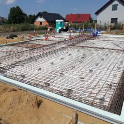 Przygotowany fundament z izolacją, zbrojeniem i instalacją ogrzewania podłogowego, widoczne rury grzewcze w białym kolorze, wystające pionowe rury instalacyjne w kolorach czerwonym i niebieskim...