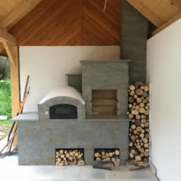 Kamienny piec chlebowy z miejscem na drewno, wykończony szarym kamieniem, pod drewnianą konstrukcją zadaszenia.