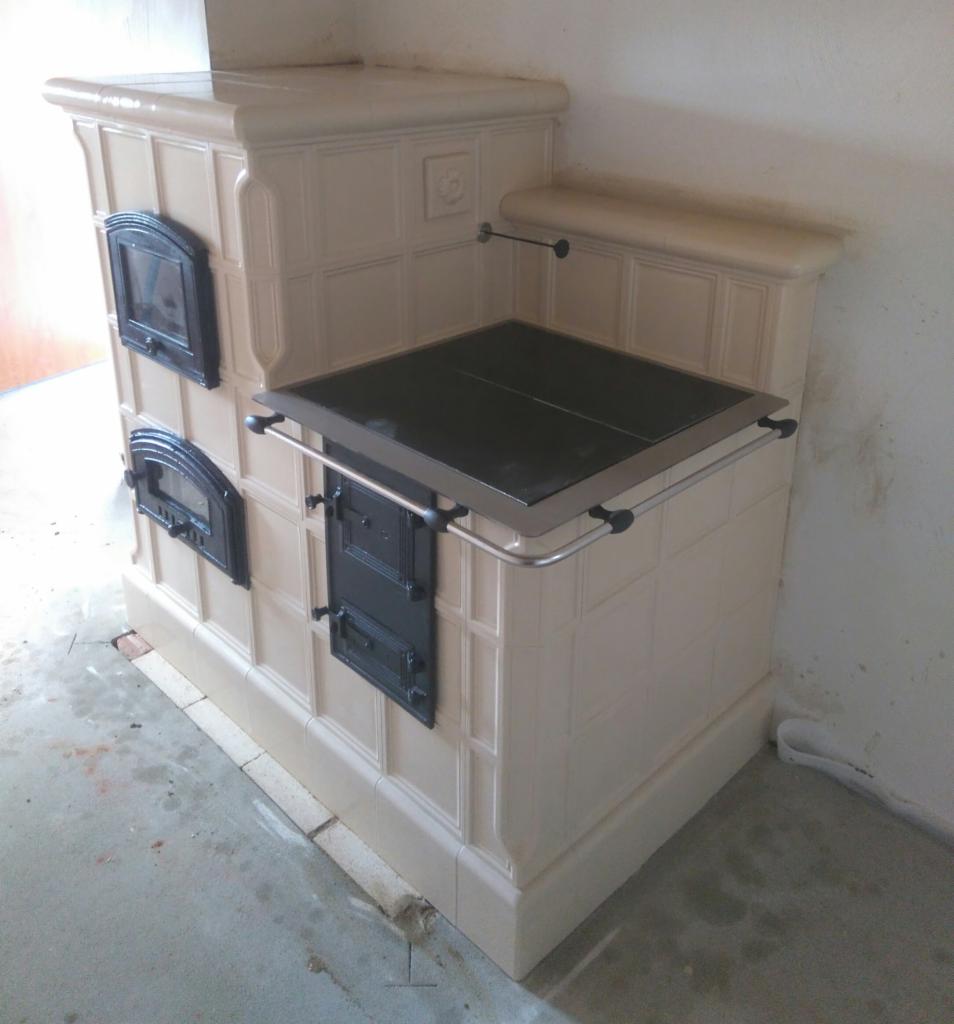 Nowy piec kaflowy w beżowej kolorystyce, z czarnymi drzwiczkami i metalowymi relingami, jeszcze nieużytkowany, z widocznymi niedoskonałościami wykończenia podłoża.