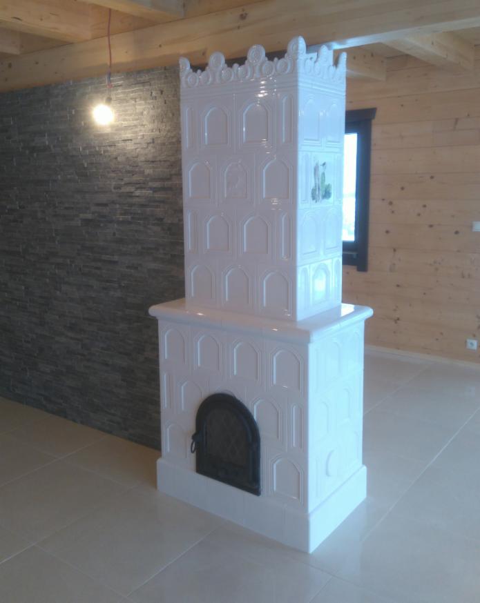 Biały, kaflowy piec z dekoracyjnymi elementami w nowoczesnym wnętrzu z drewnianą boazerią i kamienną ścianą, oświetlony wiszącą żarówką.