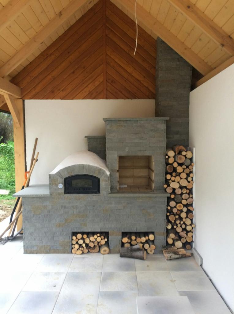 Kamienny piec chlebowy z miejscem na drewno, wykończony szarym kamieniem, pod drewnianą konstrukcją zadaszenia.