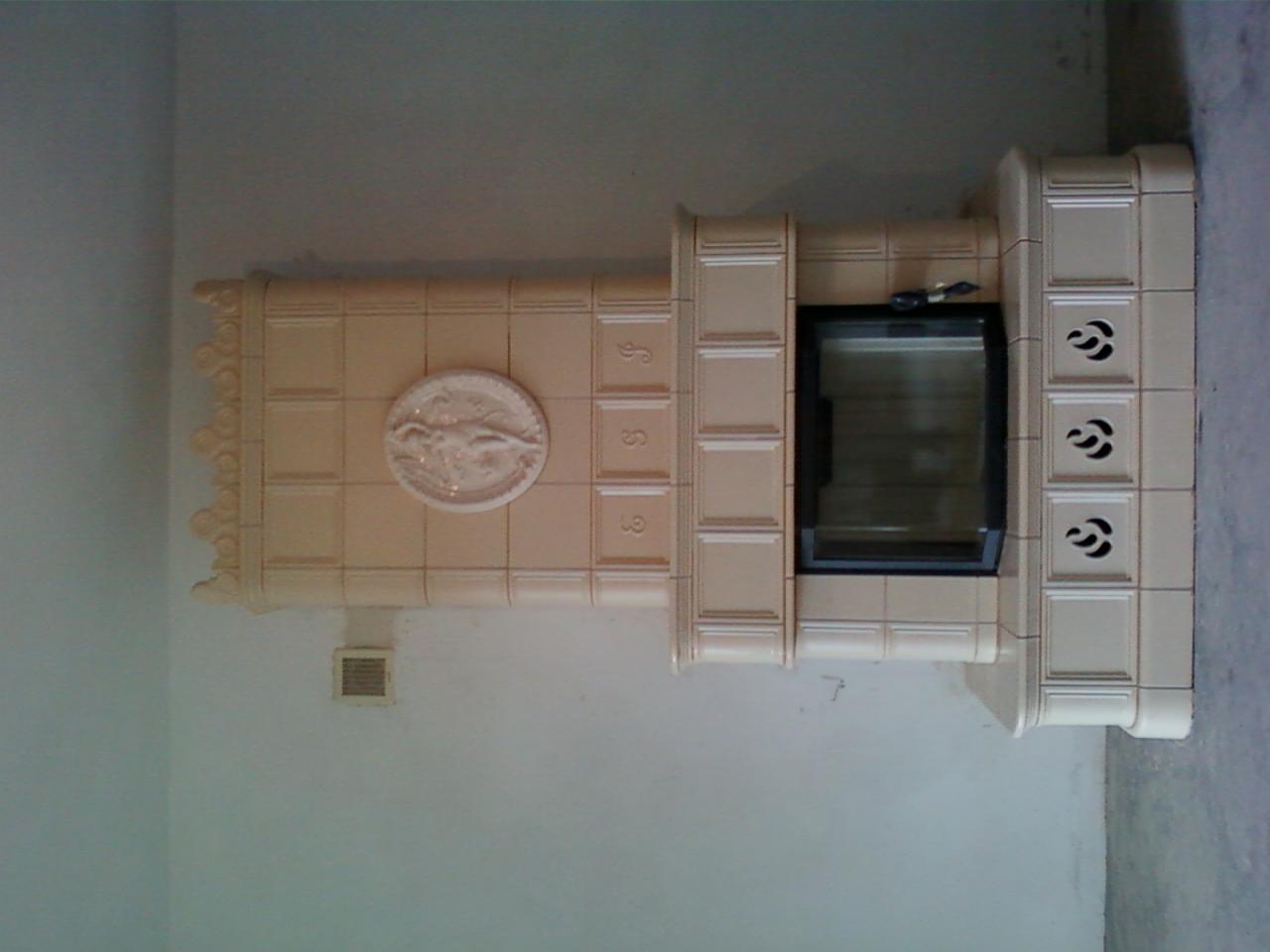 Beżowy piec kaflowy z ozdobnym medalionem i ażurowymi wstawkami, czarne drzwiczki paleniska, widoczny fragment ściany.