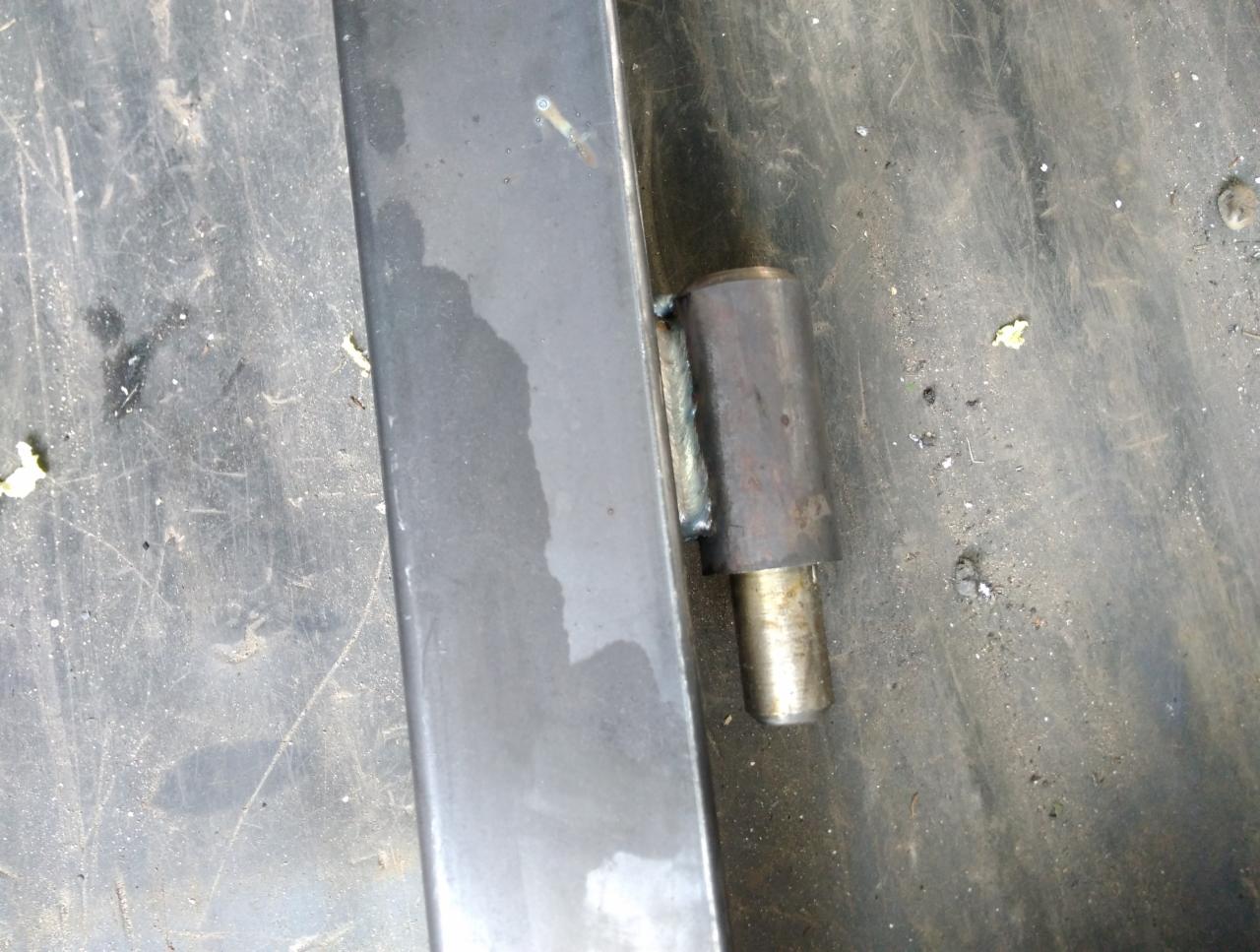Metalowy zawias przyspawany do profilu, widoczne ślady spawania na brudnym stole warsztatowym.