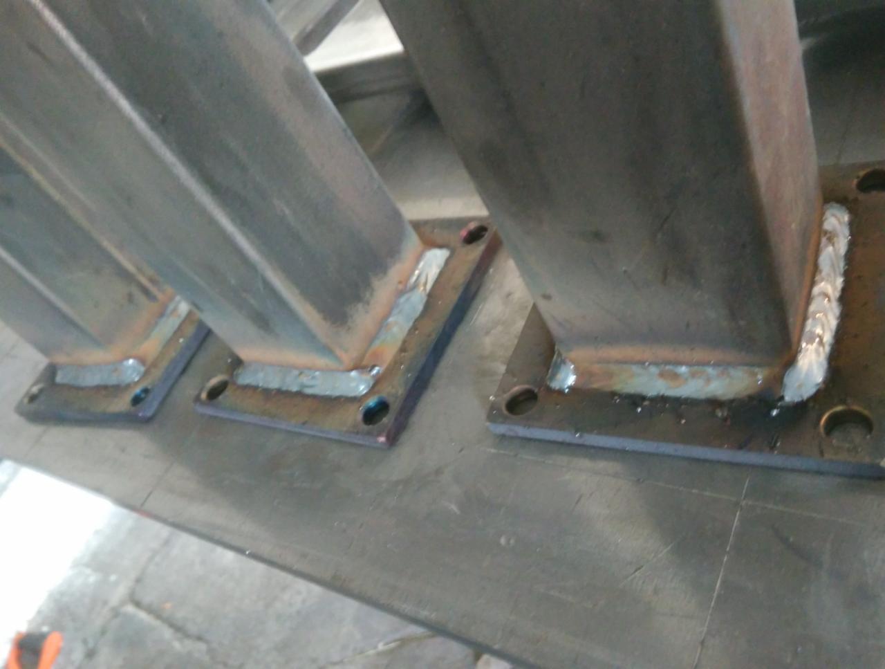 Wspawane metalowe słupki o kwadratowym przekroju, przymocowane do blachy z otworami na śruby, widoczne spoiny.