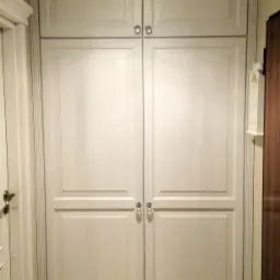 Dwudrzwiowa, sięgająca sufitu, biała szafa z ozdobnymi uchwytami i listwami pionowymi, oświetlona wiszącą lampą, umieszczona w wąskim korytarzu z drewnianą podłogą.