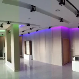 Nowoczesne wnętrze biurowe z oświetleniem LED w kolorach zielonym, niebieskim i fioletowym, z białymi panelami ściennymi i reflektorami na szynoprzewodach.