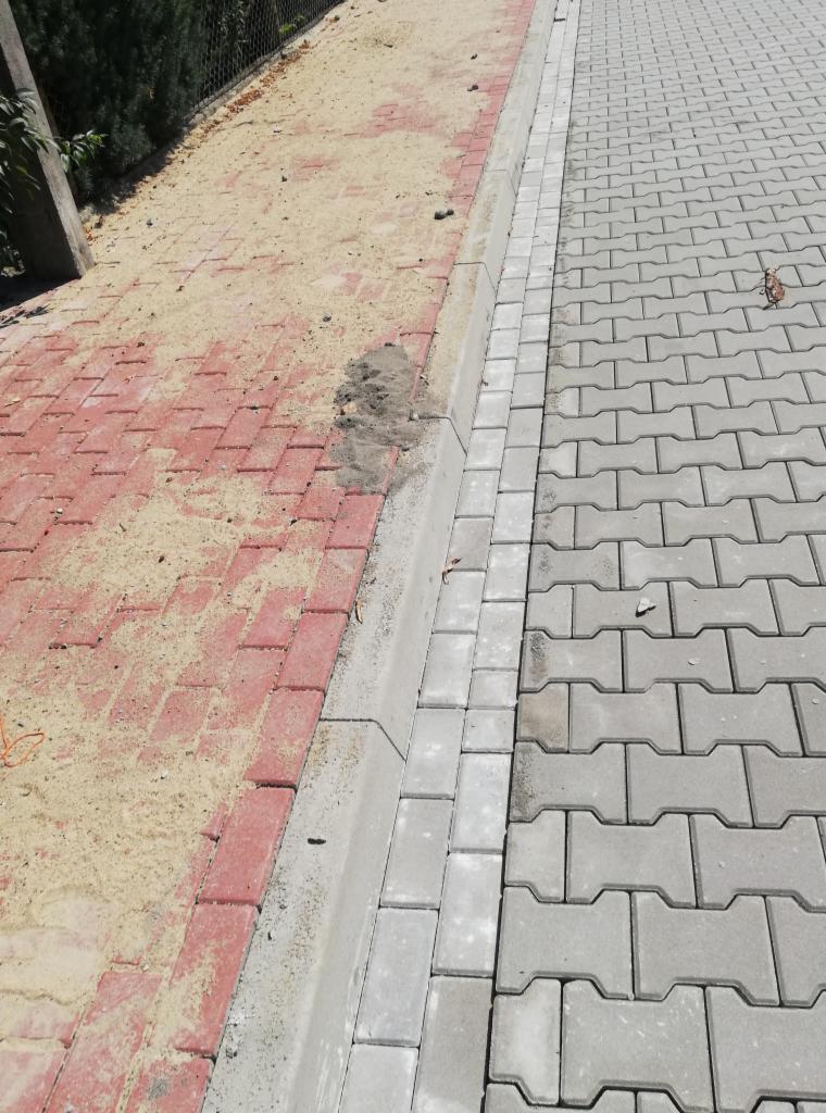 Fragment chodnika z ułożoną szarą kostką brukową w kształcie litery H, oddzieloną obrzeżem od czerwonej kostki brukowej zasypanej piaskiem, widoczne pozostałości piasku na szarej kostce.