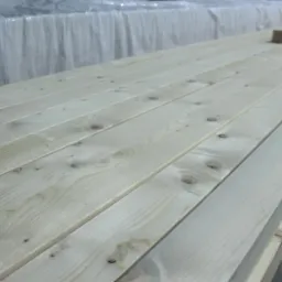 Stos jasnych, heblowanych desek sosnowych z widocznymi sękami, zabezpieczonych folią na górze.