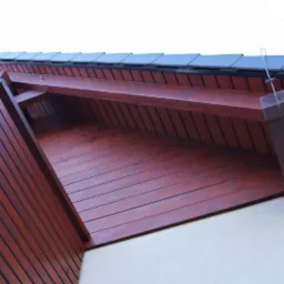 Fragment domu z drewnianą konstrukcją dachu, pomalowaną na ciemny czerwono-brązowy kolor, widoczne ciemne dachówki i fragment ściany o jasnej teksturze.