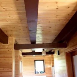 Wnętrze domu z drewnianą boazerią, sufitem z jasnych desek i ciemnymi belkami stropowymi, widoczny fragment ceglanego komina i dekoracyjny zegar.