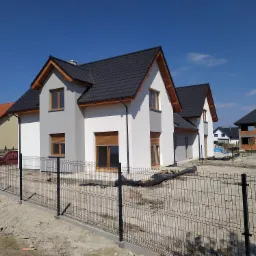 Nowe domy jednorodzinne z ciemnymi dachówkami, szarą elewacją i drewnianymi ramami okiennymi, otoczone metalowym ogrodzeniem panelowym na placu budowy.