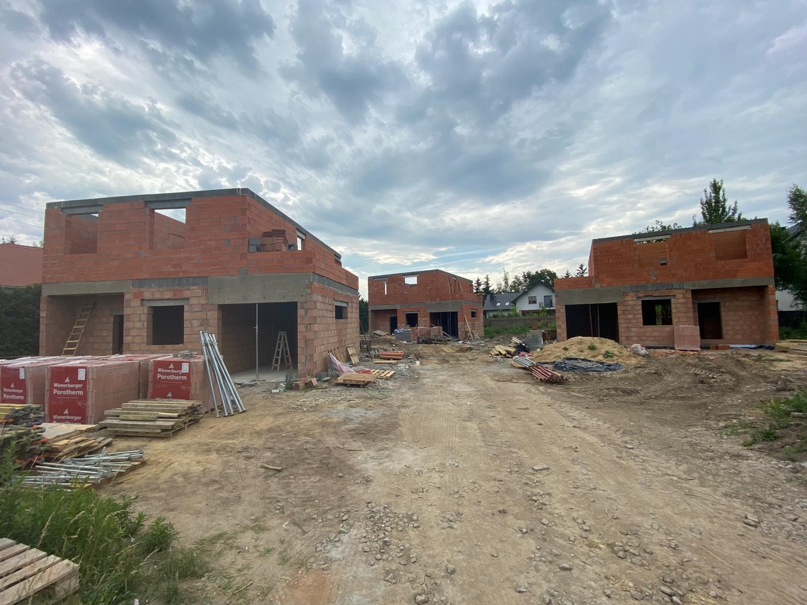 Budowa osiedla domów jednorodzinnych z czerwonej cegły Porotherm, widoczne palety z materiałem budowlanym oraz drabiny opierające się o ściany, pochmurne niebo.