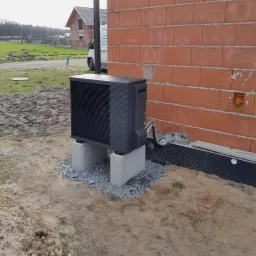 Czarna pompa ciepła zamontowana na betonowych fundamentach przy ścianie budynku w trakcie budowy. Widoczne przyłącza rur. W tle budynek w stanie surowym otwartym i samoch&oacute;d dostawczy.