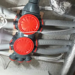 Szare rozdzielacze wentylacyjne z czerwonymi pokrywami i podłączonymi elastycznymi przewodami wentylacyjnymi, zamontowane pod sufitem.