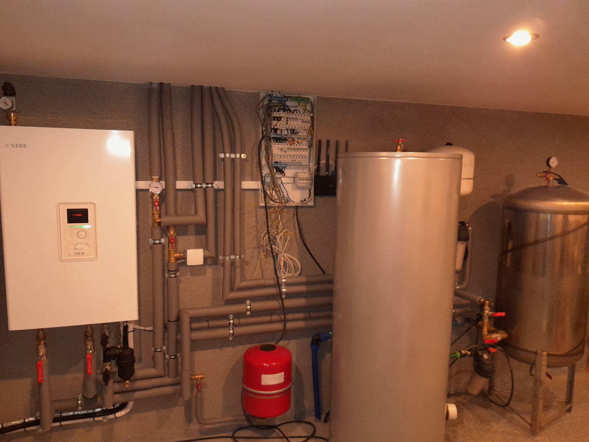 Kompletna instalacja grzewcza w kotłowni: biały piec gazowy Nibe SHB 20, dwa zbiorniki na wodę (jeden ze stali nierdzewnej na metalowym stelażu), czerwony zbiornik wyrównawczy, szare rury grzewcze...