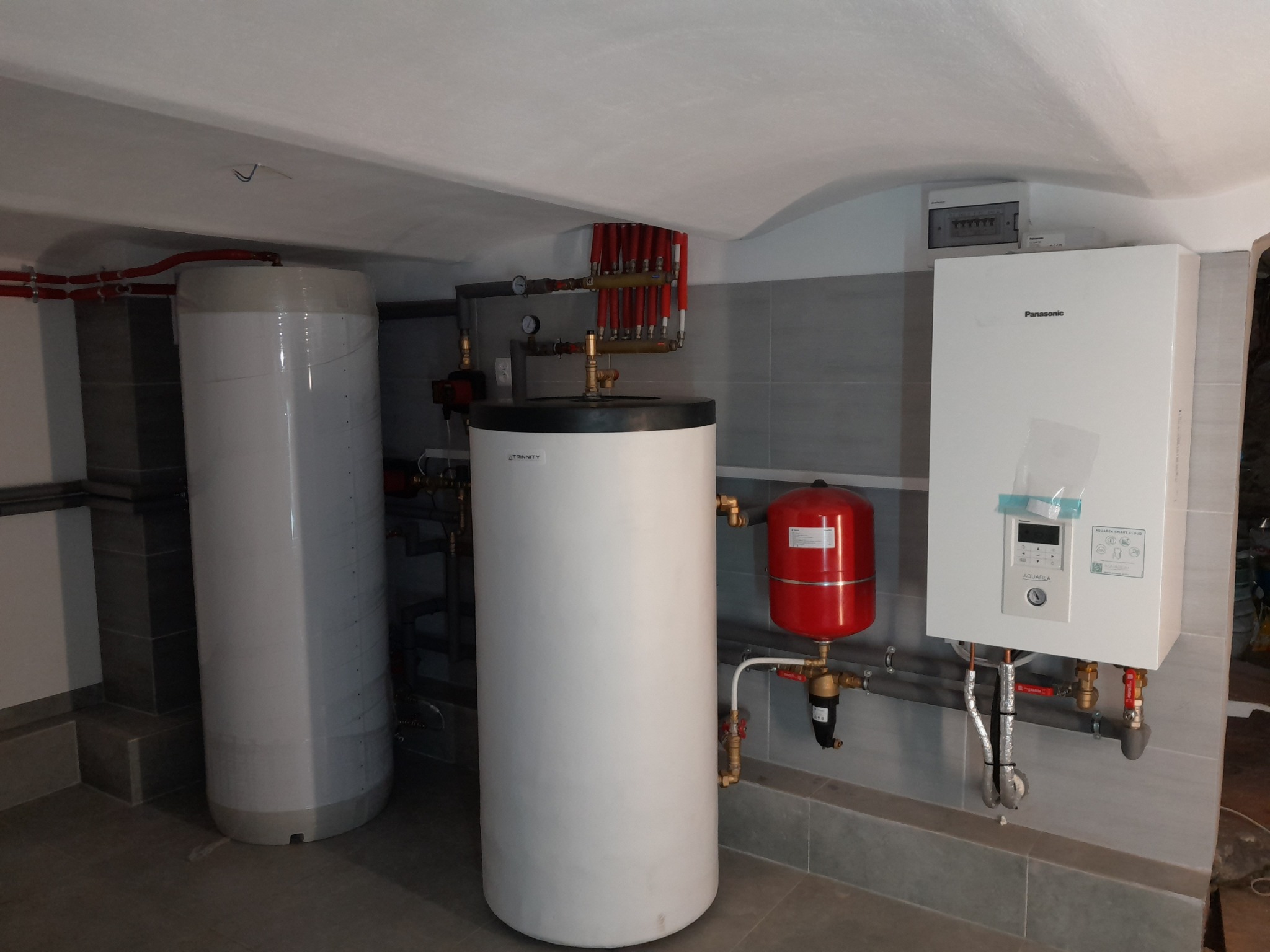 Kotłownia z dwoma białymi zasobnikami ciepłej wody, czerwonym naczyniem przeponowym i białym piecem gazowym Panasonic na tle szarych płytek.