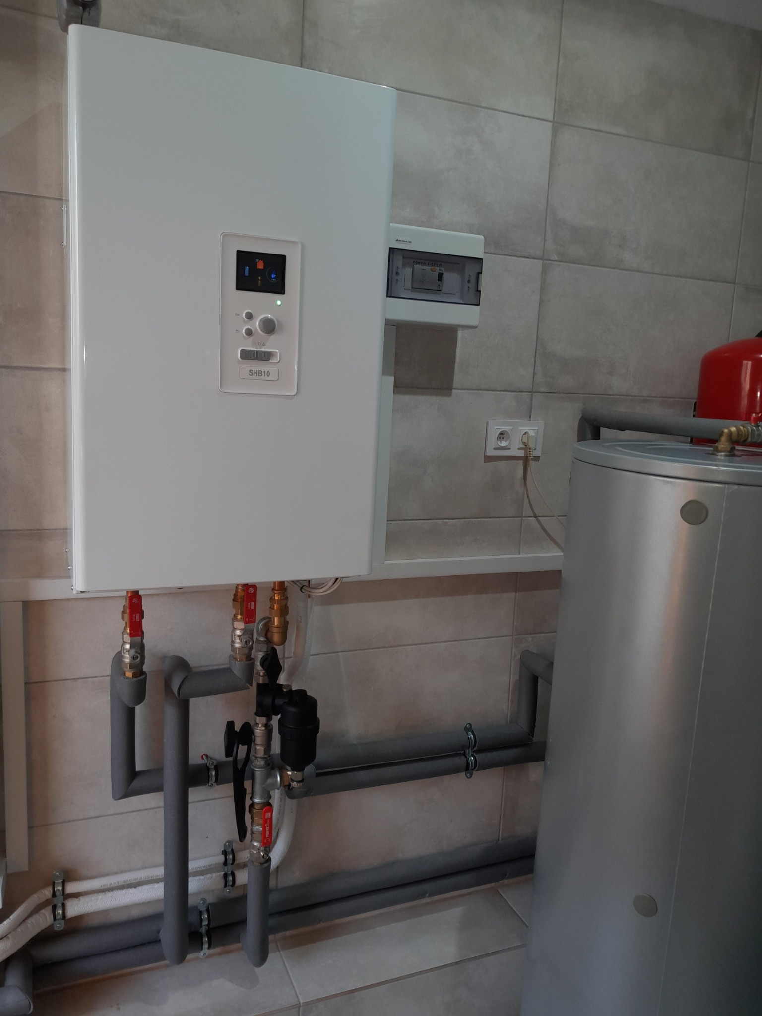 Biały piec gazowy SHB10 zamontowany na ścianie z szarymi płytkami, z widocznymi rurami, zaworami, zbiornikiem wody i skrzynką elektryczną w tle.